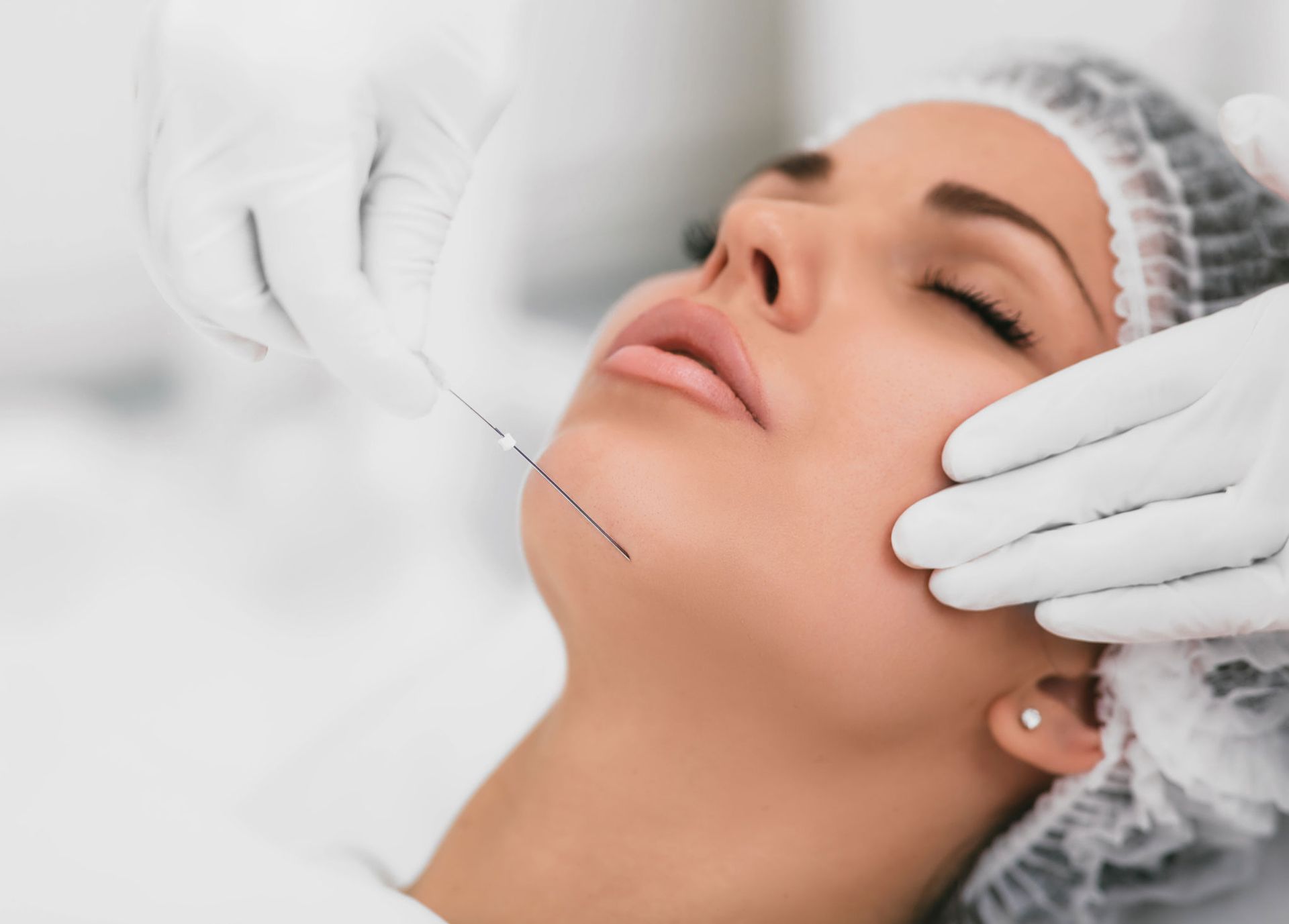 Woman receiving a cosmetic procedure on her chin; doctor's gloved hands, needle, and white setting.