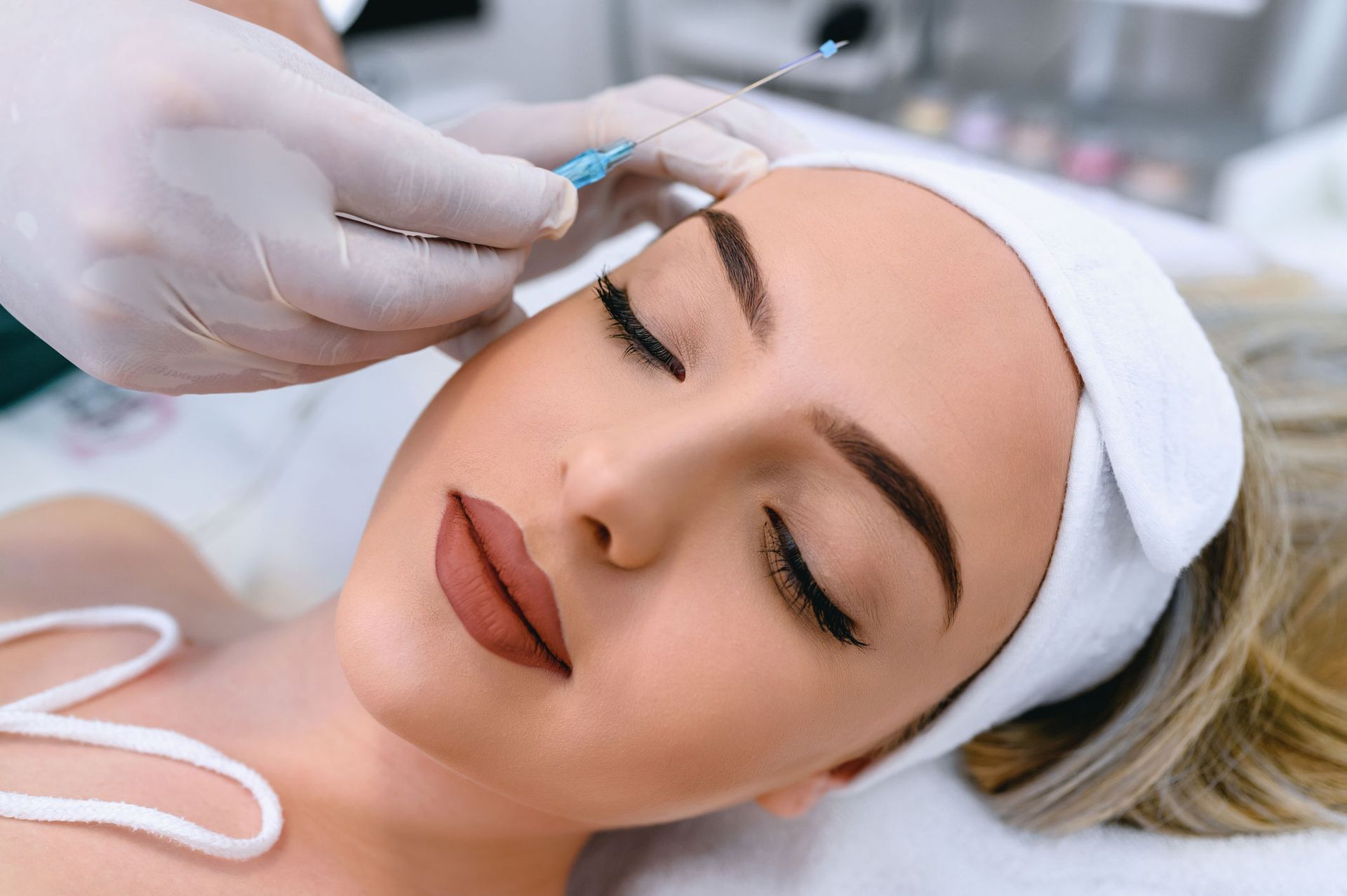 Person receiving a cosmetic procedure on the forehead. Hands with gloves hold a needle, and the person has eyes closed.