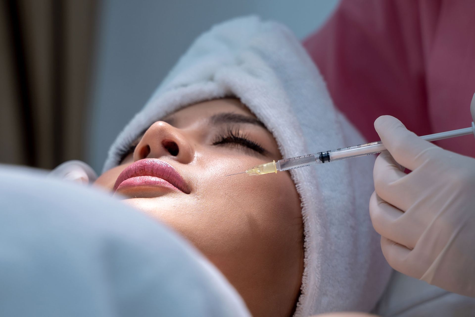 Woman receiving facial injection; close-up shows needle entering cheek, wearing white towel and pink top.