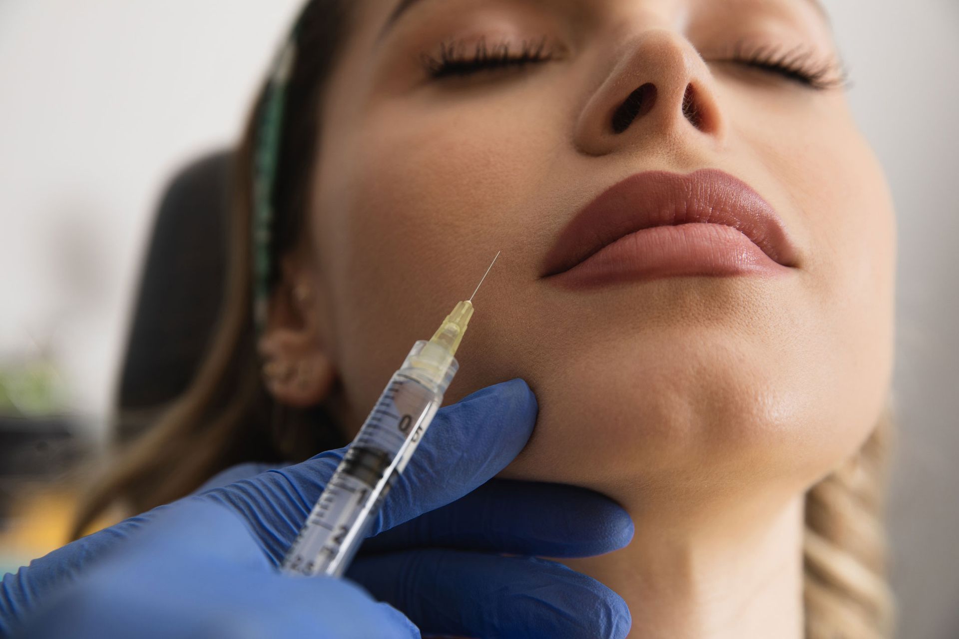 Person receiving a facial injection. A gloved hand holds a syringe near the chin of a person with eyes closed.
