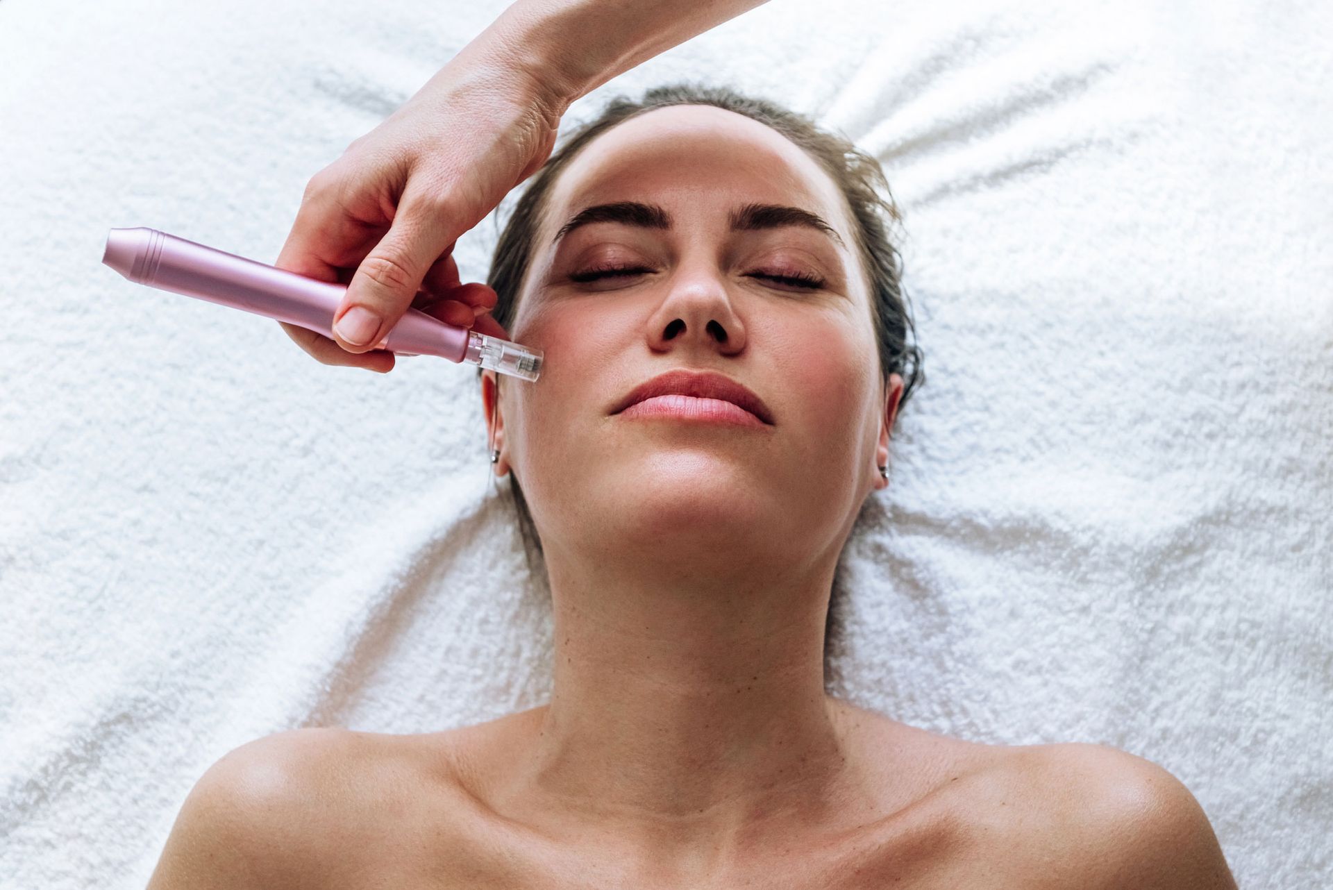 Woman receiving facial treatment, pink device held to her cheek on a white towel.