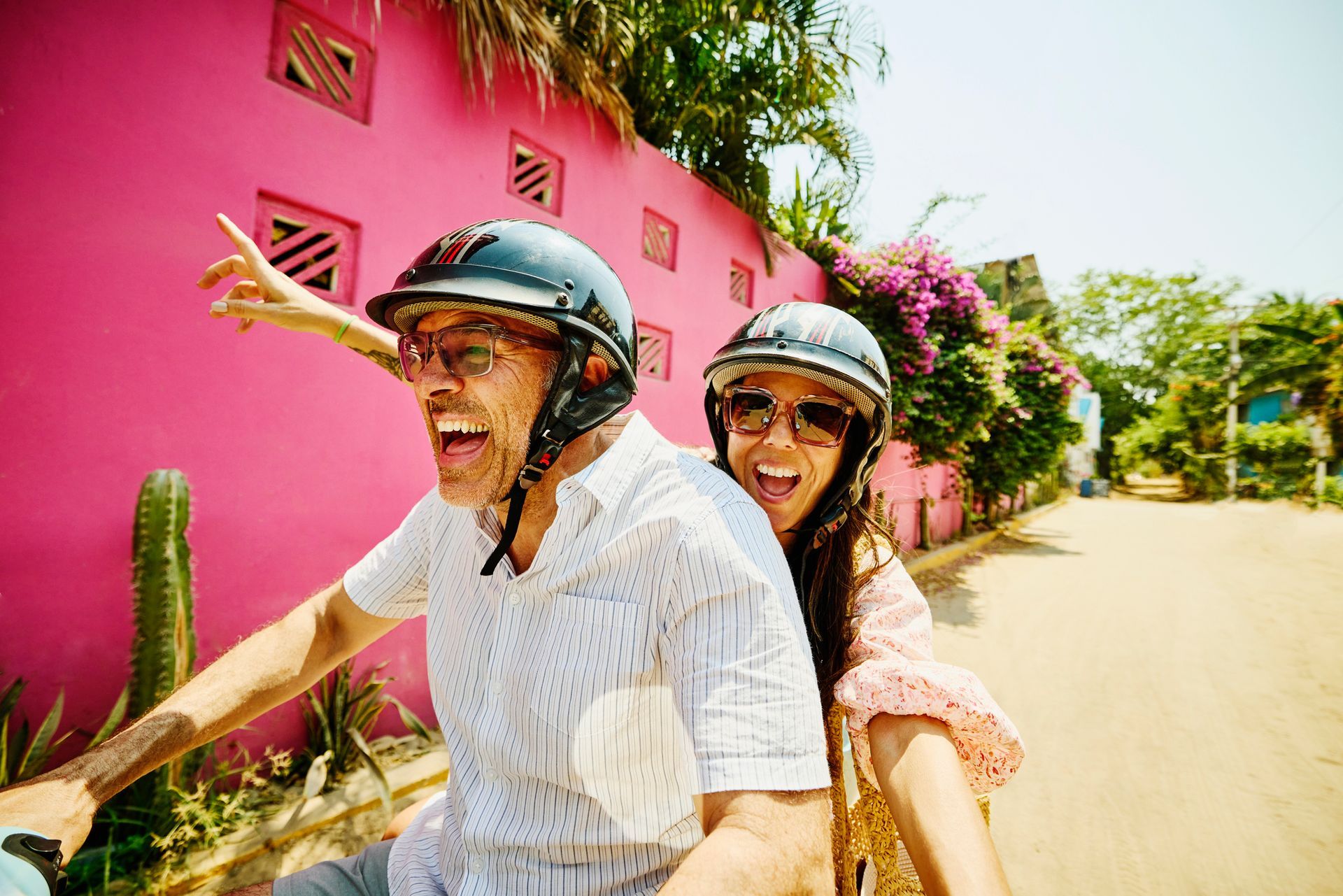 Couple on a scooter in front of a pink wall, both wearing helmets, smiling, and pointing.